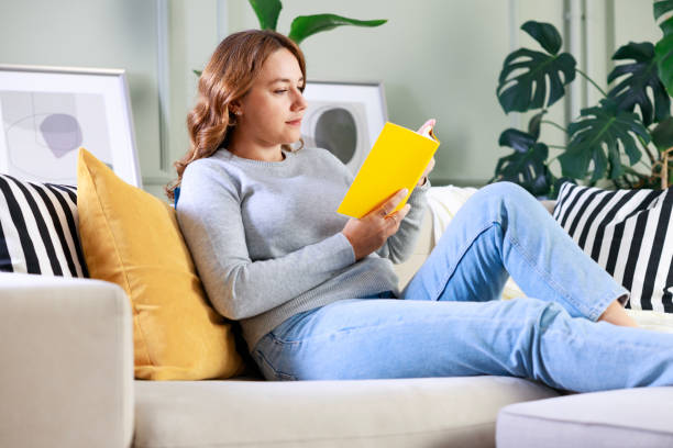 istock Young woman sitting on sofa and reading book 1748536587