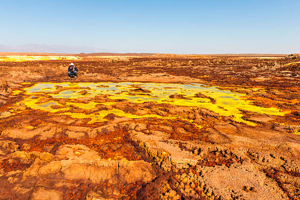 Hot Springs on Dallol Mountain "A tourist (at the distance) man sitting at the Hot Springs and geothermal / volcanic activity on Dallol Mountain. The characteristic white, yellow and red colours are the result of sulphur and potassium salts coloured by various ions. Dallol volcano is located in the Danakil Depression with the highest average temperatures on the planet. Ethiopia." dallol ethiopia stock pictures, royalty-free photos & images