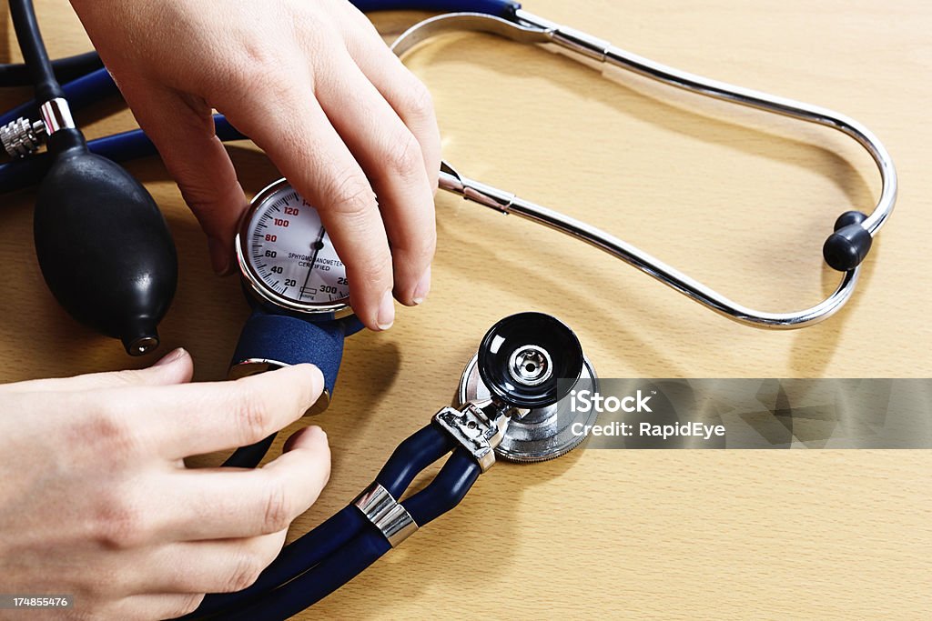 Hands holding the dial of a blood pressure gauge "Female hands hold a doctor's blood pressure gauge, or sphygmomanometer; next to it  a stethoscope sits on a wooden desk. Every medical professional's basic equipment." Blood Pressure Gauge Stock Photo