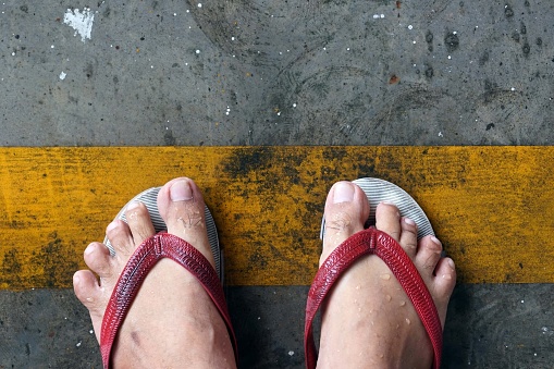 Close up of man's feet wearing red sandals stand on the yellow bar on the road in the car park.
