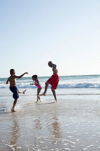 African American man (30s) playing at the beach with two children (6 years), splashing in surf.