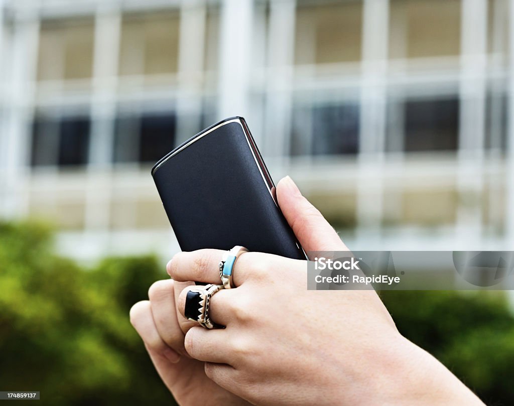 Feminine hand using smart phone outside office building A woman's hands with multiple rings busy tapping the screen of a smart phone outside an office building. Building Exterior Stock Photo