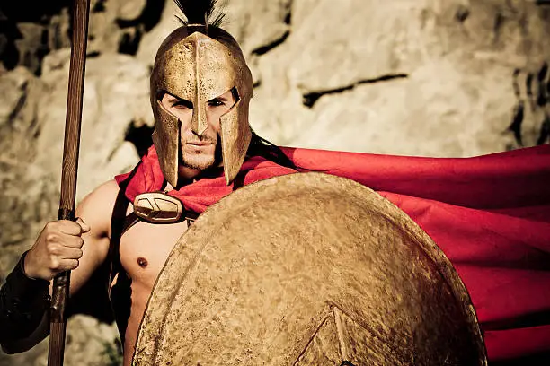 "Portrait of Spartan warrior with selfmade theater clothings and with original spartan symbol, adopted in the 420s BC, was the letter lambda (a), standing for Laconia or Lacedaemon, which was painted on the Spartans' shields and broochs,selective focus, very creative sepia - color retouching to underline the ancient time,vignetting and added noise"