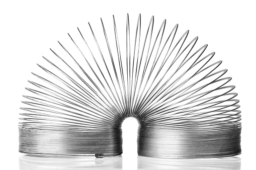 A child’s metal slinky toy is isolated on a white background with a clipping path. The metal slinky is folded over in half, which is the position it is put into to start moving down stairs. It reflects into the shiny white background that it is sitting on.