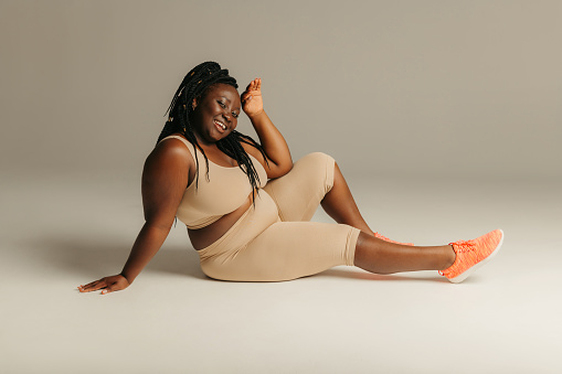 Joyful plus curvy African woman in sportswear radiating self-love while sitting against studio background