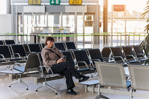 Adult traveler woman sits at airport terminal awaits boarding a flight for departure while uses smartphone with free wi fi. Concept of people sharing informations with new technology while traveling