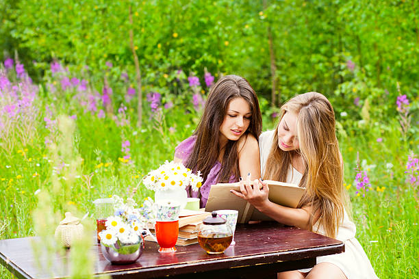 Two young women in a summer garden reading a book and drinking tea.