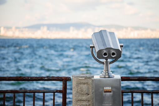 Public binoculars on the seaside promenade overlooking a distant city.