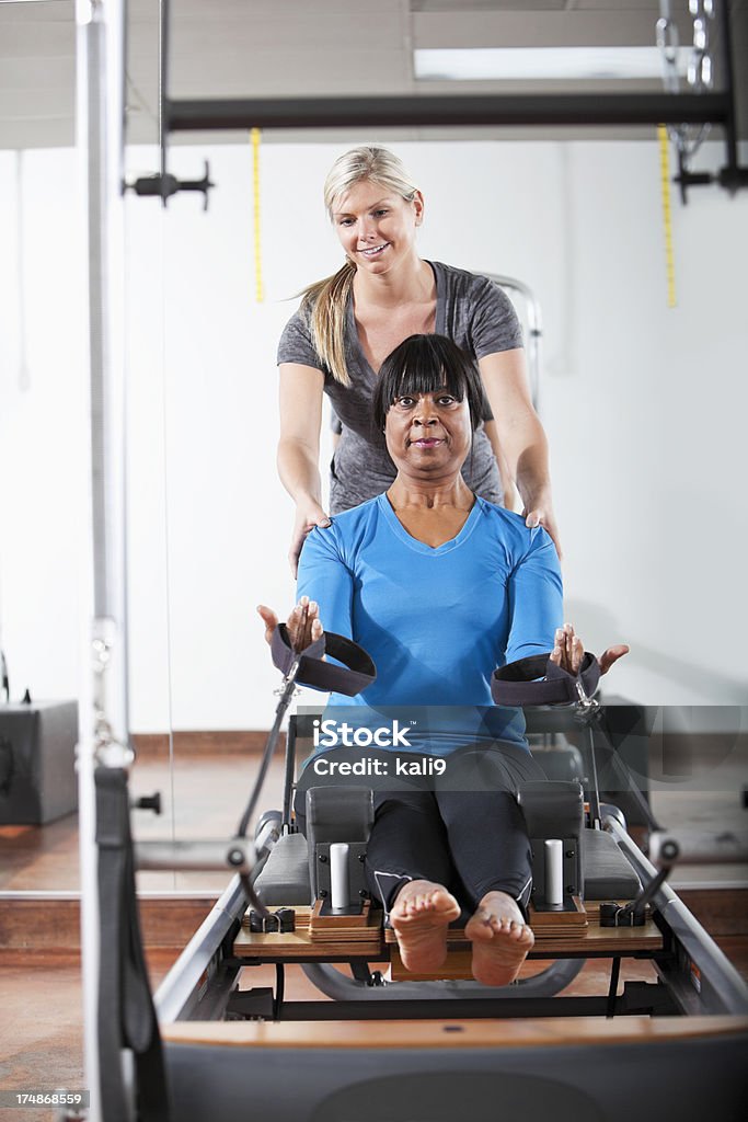 Personal trainer helping woman on pilates reformer Instructor (20s) helping African American woman (60s) on pilates reformer. 20-29 Years Stock Photo