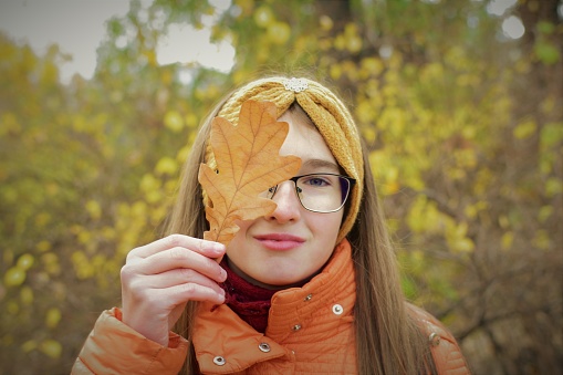 A beautiful girl with glasses covers half of her face with an autumn oak leaf. Bokeh in the background.