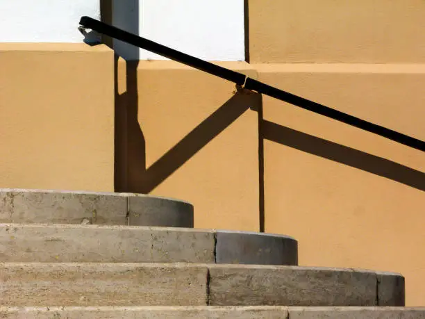 curved exterior concrete stair closeup. risers, treads. yellow stucco wall elevation. black iron wall attached handrail. strong sunlight and shadows. summer scene. architecture and design concept.