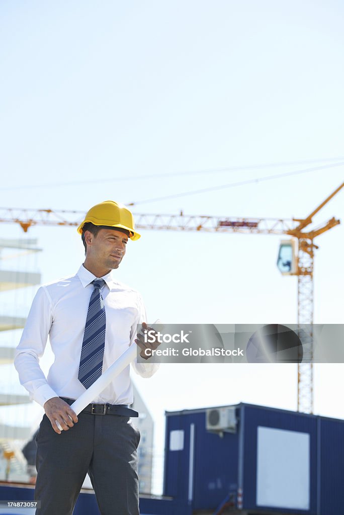 Friendly and focused An architect stands on a construction sight with his building plans in hand Adult Stock Photo