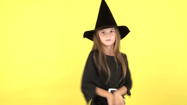 A little girl in a Halloween witch costume dances and shows emotions on a yellow studio background