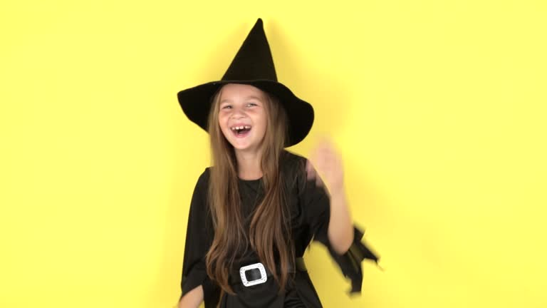 Happy Halloween concept. Portrait of little girl in witch costume raises hands scares and looking at camera, posing isolated on yellow color background wall in studio.