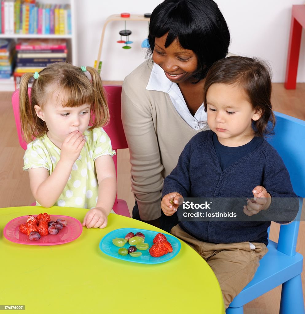 Feliz professor/cuidador supervisionando menino e menina no horário das refeições - Foto de stock de Criança pequena royalty-free