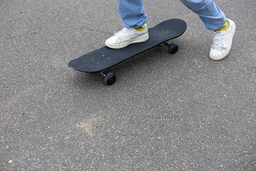 The legs of a child in jeans and white sneakers stand on a skateboard on an asphalt road. A child rides a skateboard on the street. Feet push off the asphalt on a skateboard..