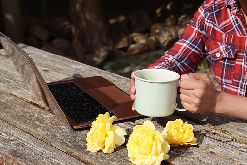 Women working on the computer laptop with an Enamel mug of coffee. Woman sit on the old wooden table working online business in the garden at home. Lifestyle photo.