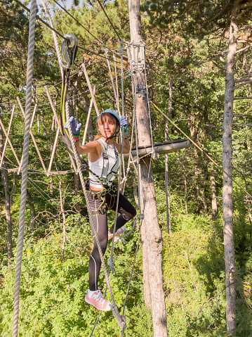A teenager Girl walks along a rope bridge between trees in an amusement park in safety gear and a helmet. Rope park in wood forest.High-altitude climbing training of child on adventure track, equipped with safety straps and protective helmet.