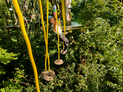 High-altitude climbing training on adventure track, equipped with safety straps.A people walks a rope bridge between trees in an amusement park in safety gear and a helmet. Rope park in wood forest.A group of people goes on hinged trail in extreme rope Park in summer forest.