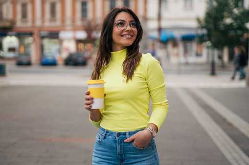 Portrait of beautiful young woman in yellow turtle neck in the street holding cup of coffee.