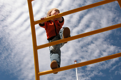 A 2 year old is climbing on a yellow monkeybar at a playground outside