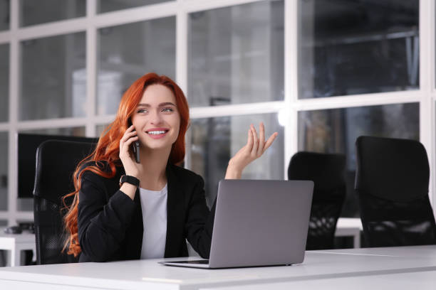 istock Happy woman talking on smartphone while working with laptop at white desk in office 1749079028
