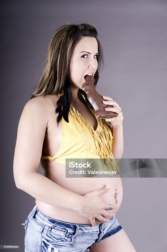 Pregnant woman eating chocolate bunny, holding belly. "Vertical studio shot on grey of pregnant woman with one hand on belly, other holding doomed Easter bunny." One Woman Only Stock Photo