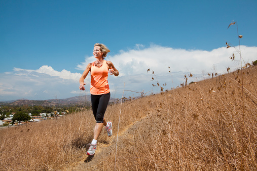 Attractive Mature Woman is determined to stay fit and healthy. Outdoors shot of a nature trail with white clouds on a bright blue sky. Fit, mature woman in her 50s is running and jogging on the dirt trail. Woman is athletic built and is body conscious. She lives a healthy lifestyle and exercises regularly.   