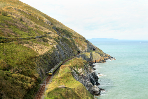 a commuter train can be seen on the new railway tunnel and track on coastal railway Wicklow Ireland - the famous coastal railway between Bray and Greystones near Dublin, Ireland. The original coast railway had been badly eroded and was replaced by a new tunnel system devised by Isambard Kingdom Brunel, and hampered by the very tough granite cliffs. The current railway hosts the Dublin Area Rapid Transit (DART) commuter light railway. Dublin Bay and Howth peninsula can be seen in the background. The famous scenic coastal path can be seen on the cliffs above the railway line.