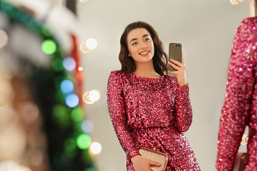 Beautiful young woman trying on stylish pink sequin dress and taking selfie near mirror in boutique. Party outfit