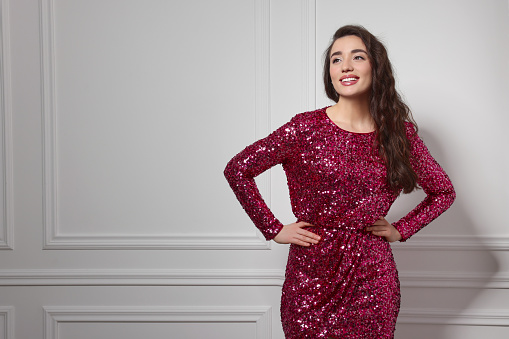 Beautiful young woman in stylish pink sequin dress near white wall indoors, space for text. Party outfit