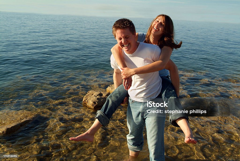 Middle aged couple playing on the beach Horizontal image of a playful young couple having fun near the water's edge. She is riding on his back and the man is acting as if he is not strong enough to carry her. Both have cute expressions. The water is a beautiful blue and fades into the blue sky.  Adults Only Stock Photo