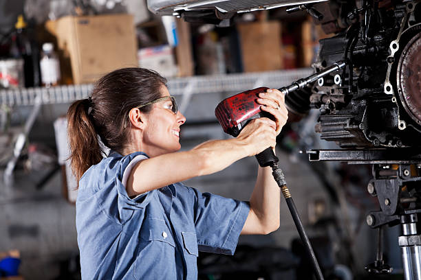 Female auto mechanic working on car transmission Female auto mechanic with a power tool fixing a car's transmission. She is wearing safety glasses and a denim short-sleeved shirt. She is holding the power tool with both hands. car mechanic top view stock pictures, royalty-free photos & images