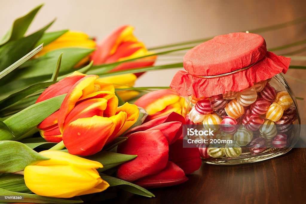 Colorful tulips and candies on a wooden surface A bunch of red and yellow tulips with leaves lay on a wood grain surface to the left of a jar that holds striped candies.  The tulips include a solid red and a solid yellow blossom; the rest are red with yellow-edged petals.  Center of focus is the small jar, which has a red fabric cover tied on with a gold strand.  The candies are small and round with white stripes, and include green, red, pink, yellow and purple in the clear glass jar.  Light glows softly off the wood grain in the background. Candy Stock Photo