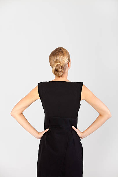 Businesswoman in a black dress faces away holding hands on hips on gray background. Studio shot