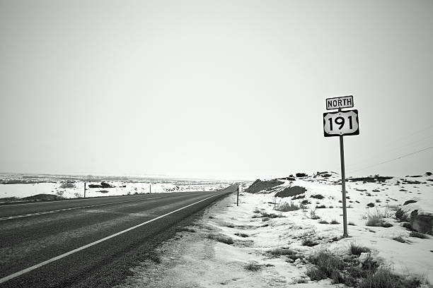 winter road trip road sign north 191 and asphalt cut through the snow covered desert landscape.  horizontal wide angle composition taken in moab utah.  overcast skies caste the landscape in intense grays and blacks with retro black and white color treatment. sonoran desert desert badlands mesa stock pictures, royalty-free photos & images