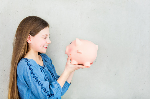 Portrait of a happy girl with a piggy bank. Lots of copyspace and room for text on grey wall