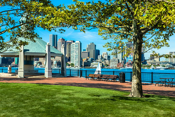 Cityscape from the East Boston Pier's Park A brick walkway in East Boston's Pier's Park invites you to stroll past the lush green grass along the black wrought-iron railing by the waterfront. Relax on the benches, or enjoy lunch at the picnic table while gazing out across the Boston Harbor to see the sailboats and downtown's numerous buildings. Or enjoy the sights from the strategically placed, beautiful pavilion with its conical-shaped green roof. HDR Image. east boston stock pictures, royalty-free photos & images