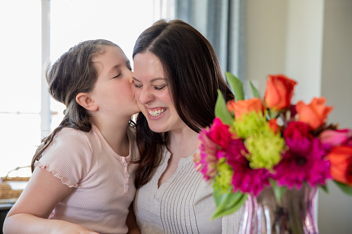 A young girl kisses her mother on the cheek while a vase of flowers sits on the counter