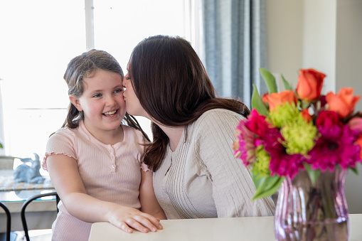 A mid adult mother kisses her young daughter on the cheek while a vase of flowers sits on the counter