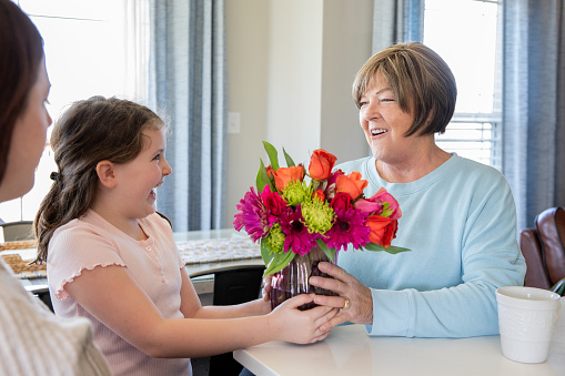 A young granddaughter presents her grandmother with a vase of flowers while her mother looks on