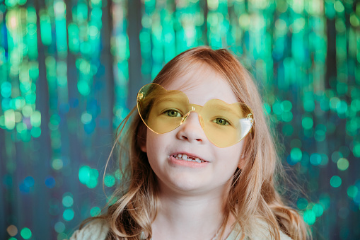 A young girl wears heart-shaped glasses and smiles at the camera in front of a shiny backdrop