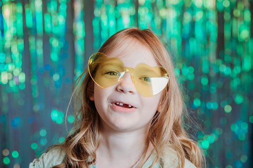 With a shiny backdrop, a young girl sports heart-shaped glasses and beams at the camera