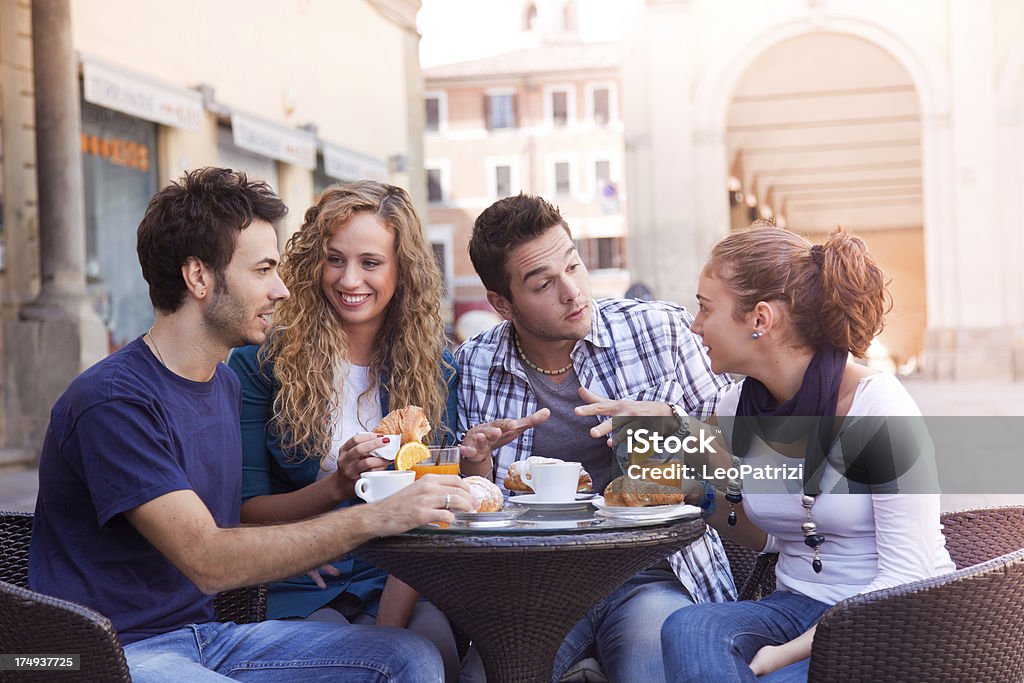 Happy friends having breakfast outdoor Four young people sit around a small table in the sunlight.  Two are women and two are men, seated as two couples.  There are two coffee mugs and two orange drinks in front of them, and several buildings behind them. 20-24 Years Stock Photo