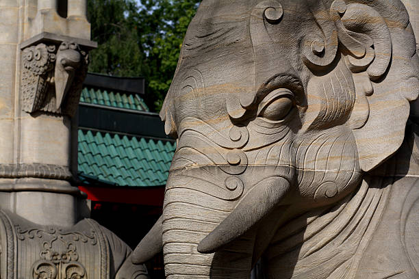 Thai man is getting up on elephant. Asian elephant is posing front leg as climbing aid. Scene is close to King Naresuan monument in Lampang. Elephant is standing on small square near monument. In background is articial ancient looking wall for upcoming historical event around monument following day. Part of a series of photos