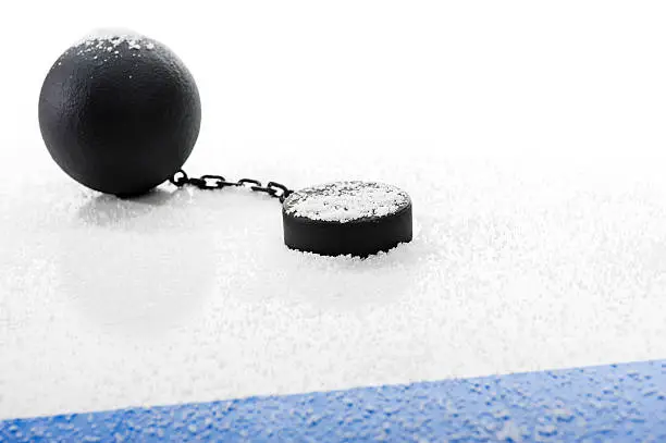 Concept of the NHL Lockout, with an idle hockey puck attached to a ball and chain covered in snow next to the Blue Line agaist a white background