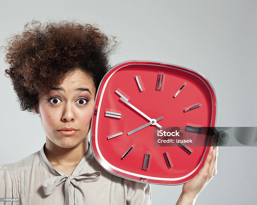 Afro Girl with Clock Portrait of teenaged afro girl holding a big red clock next to her head, staring at the camera. Studio shot, grey background. Clock Stock Photo