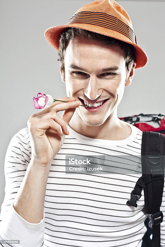 Happy backpacker Portrait of young adult man wearing hat and backpack smiling at the camera with small flower in his teeth. Studio shot, grey background. Young Men Stock Photo
