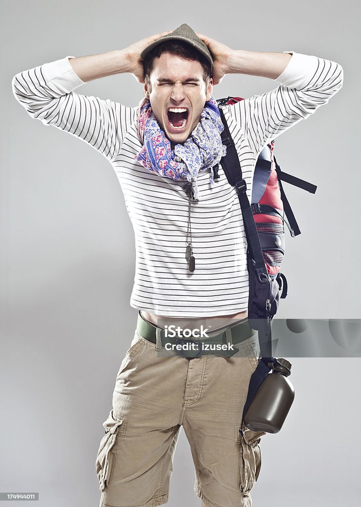 Angry traveller Portrait of angry young backpacker with hands behind head screaming at the camera. Studio shot, grey background. Young Men Stock Photo