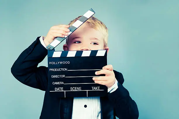 Boy with blond hair hold an open Hollywood slate board. He is looking to the side and looks quite happy. The boy is wearing a tuxedo.